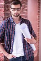 Architect with blueprint. Confident young man in glasses holding blueprint and looking at camera while standing indoors photo