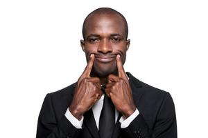 Forced smile. Serious young African man in formalwear making a smile by his fingers while standing isolated on white background photo