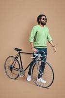 Full length of young African man in casual clothing walking with bicycle while standing against brown background photo