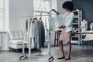 Full length of attractive young African woman looking at sleeveless shirt and touching it with hand while standing in workshop photo