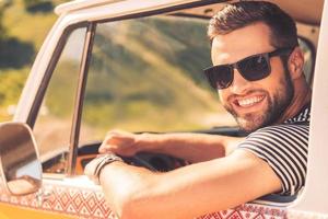 Enjoying his road trip. Cheerful young man smiling at camera and holding hand on steering wheel while sitting inside of his minivan photo