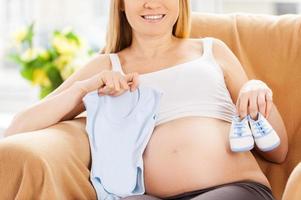 Expecting baby boy. Beautiful pregnant woman sitting on the chair and showing baby clothes photo