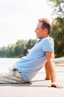 Enjoying doing nothing. Side view of cheerful mature man looking away and smiling while sitting at the quayside photo