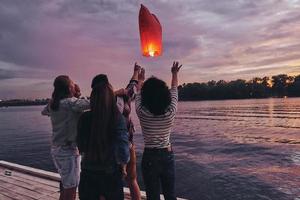 Friends forever. Group of young people in casual wear letting go the sky lantern while standing on the pier photo