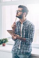 In search of inspiration. Thoughtful young man holding notebook and touching his chin with pencil while standing in front of the window photo