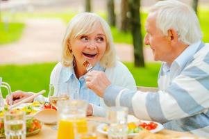 Try my meal Senior man feeding his cheerful wife with fresh salad while both sitting at the dining table outdoors photo