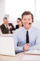 Always ready to help. Handsome young man in headset looking at camera and smiling while people working on background photo