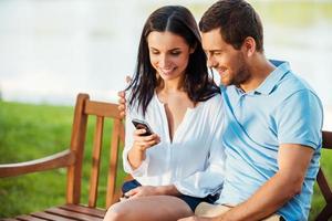 Looking their common photos together. Cheerful young loving couple sitting on the bench and looking at mobile phone together