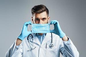 joven seguro de sí mismo con bata blanca de laboratorio ajustando la máscara protectora mientras se enfrenta a un fondo gris foto