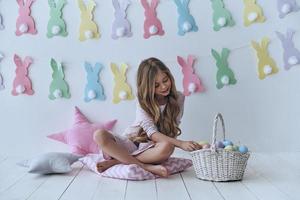 recolectando huevos de pascua. linda niñita poniendo un huevo de pascua en la canasta y sonriendo mientras se sienta en la almohada con decoración en el fondo foto