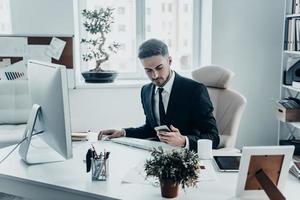 Receiving important messages.  Serious young man in formalwear holding smart phone and looking at it while sitting at the office desk photo