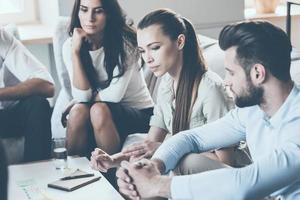 Thinking about solution. Group of young business people discussing something while sitting together and looking at paper laying on desk photo