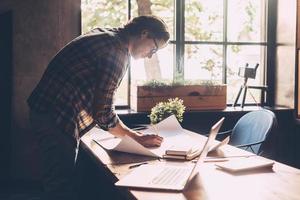 Concentrated at work. Confident young man in casual wear sketching on blueprint while standing near wooden desk in creative office photo