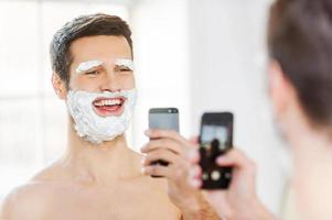 Morning fun. Rear view of playful young man with shaving cream on his face making selfie and smiling while standing in front of the mirror photo