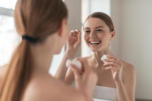 Make it a habit. Over the shoulder view of young beautiful woman holding dental floss and smiling while looking in the mirror photo