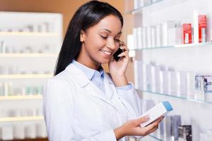 Advising a proper medicine. Beautiful young African woman in lab coat holding container with some medicine and talking on the mobile phone while standing in drugstore photo