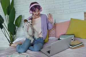 alegre adolescente usando laptop y saludando con la mano mientras se sienta en la cama con su perro foto
