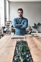 Young and successful. Handsome young man looking a camera and keeping arms crossed while standing in the creative working space photo