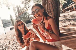 Refreshing. Two attractive young women smiling and eating watermelon while sitting on the beach photo