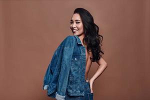 Pure feminine beauty. Attractive young woman carrying on shoulders denim jacket and smiling while standing against grey background