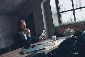 Handsome young man in smart casual wear reading a newspaper while sitting at his desk in office photo