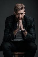 Perfect man. Beautiful young man keeping hands clasped and looking at camera while sitting against grey background photo