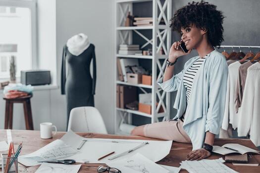 hablando de moda bella joven africana hablando por teléfono inteligente y mirando hacia otro lado mientras se sienta en el escritorio de su taller foto