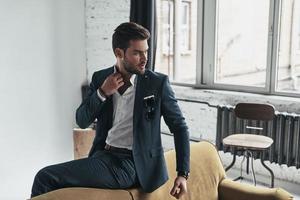 True elegance. Handsome young man in full suit adjusting his shirt and looking away while sitting on the sofa photo