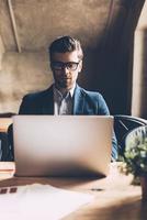 Man working on laptop. Confident young man in smart casual wear working on laptop while sitting at his working place in office photo