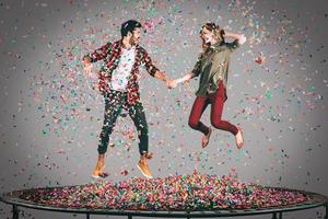 Carefree fun. Mid-air shot of beautiful young cheerful couple holding hands while jumping on trampoline together with confetti all around them photo