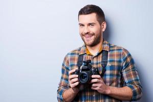 Expert in photography. Portrait of confident young man in shirt holding camera while standing against grey backgroundExpert in photography. Portrait of confident young man in shirt holding camera while standing against grey background photo