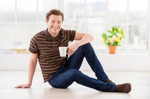 Relaxing at home. Handsome young man holding a cup and smiling at camera while sitting on the floor at his apartment photo