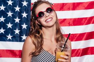 chica americana. joven alegre sosteniendo una taza de jugo y usando gafas de sol mientras se enfrenta a la bandera nacional estadounidense foto
