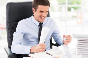 enfermo y cansado. joven furioso con camisa y corbata sosteniendo papeles en las manos y gritando mientras estaba sentado en su lugar de trabajo foto