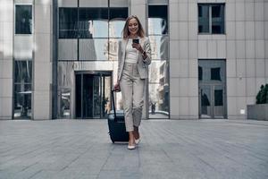 Always ready to help. Full length of young woman in suit pulling luggage and smiling while walking outdoors photo