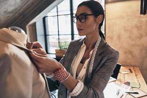 Her design is perfect. Serious young woman in eyewear adjusting a collar of the jacket on mannequin while standing in her workshop photo