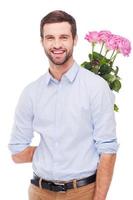 Surprise for her. Handsome young man hiding a bouquet with pink roses behind back and smiling while standing isolated on white background photo