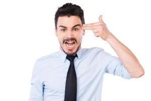 día demasiado estresante. joven frustrado con camisa y corbata gesticulando con una pistola cerca de la cabeza y manteniendo la boca abierta mientras se enfrenta a un fondo blanco foto