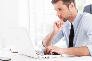 Concentrated on work. Side view of thoughtful young man in formalwear looking at laptop and holding hand on chin while sitting at his working place photo