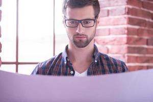 Young and confident architect. Handsome young man in glasses examining blueprint while standing near the window photo