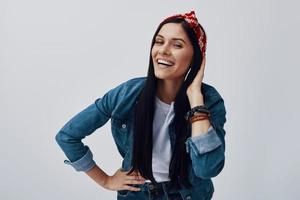 mujer joven funky en bandana mirando a la cámara y sonriendo mientras está de pie contra el fondo gris foto