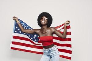 mujer africana joven feliz que lleva la bandera americana y sonriendo mientras está de pie contra el fondo gris foto