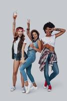 Full length of three attractive young women drinking refreshing cocktail and smiling while standing against grey background photo