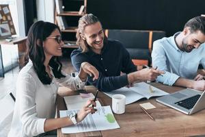 Discussing risky ideas. Group of young modern people in smart casual wear pointing at laptop and smiling while sitting in the creative office photo