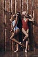 Summer joy. Full length of two attractive young women in swimwear smiling while posing against the wooden wall outdoors photo