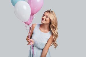 princesa feliz atractiva mujer joven sosteniendo globos y mirando a la cámara con una sonrisa mientras se enfrenta a un fondo gris foto