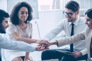 New achievements are welcome  Close-up part of top view of group of four young people holding hands and showing their unity with smile while sitting at office photo