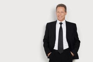 Confident in himself. Cheerful mature man in formalwear holding hands in pockets and smiling while standing against grey background photo