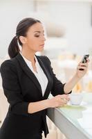 Checking business messages. Confident young woman in formalwear drinking coffee and looking at her mobile phone while leaning at bar counter photo