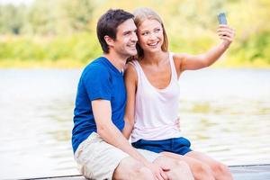 Catching great moments of life. Beautiful young loving couple sitting at the quayside and making selfie by smart phone photo